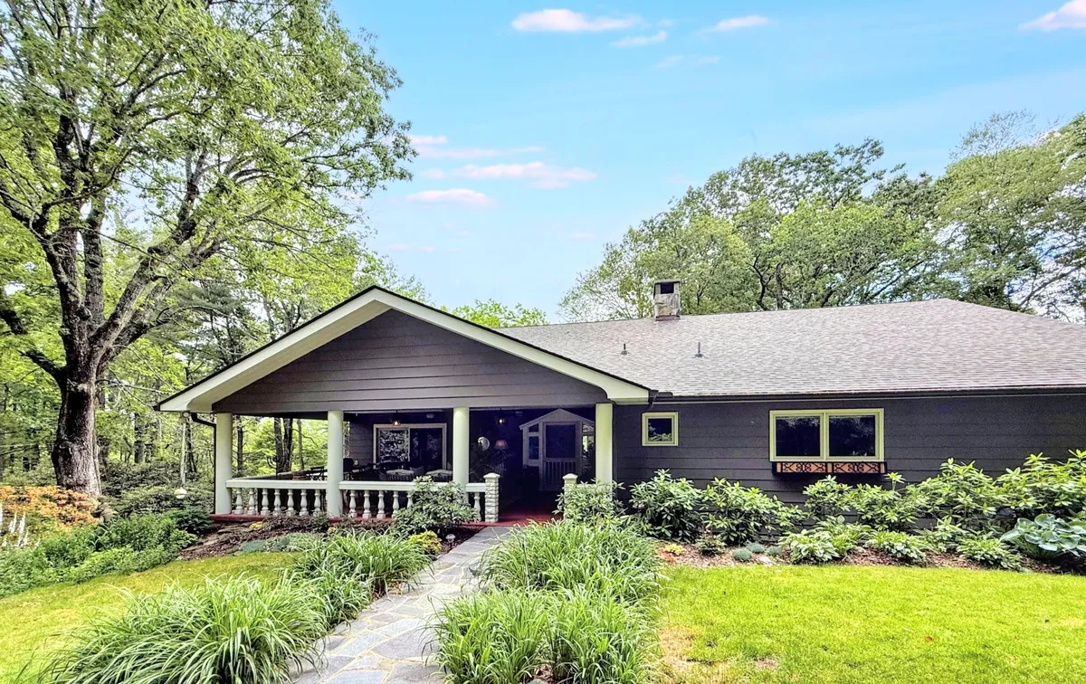 The image showcases the estate's inviting front porch framed by lush greenery, a charming stone pathway leading to the entrance, and expansive windows that hint at warm, light-filled interiors. The tranquil setting is enhanced by sunset views and seasonal touches, creating an atmosphere of peaceful retreat.