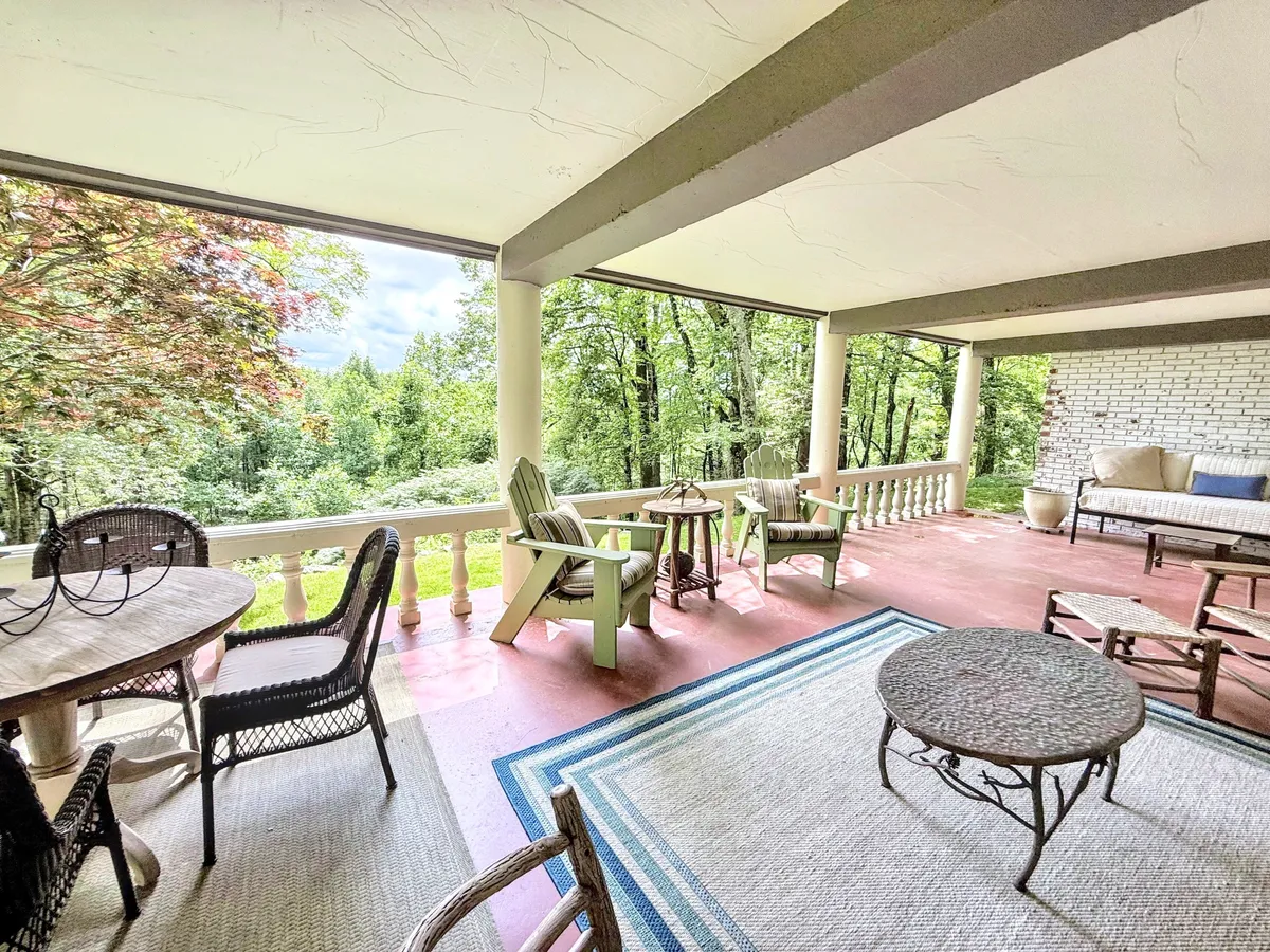 An outdoor dining and lounge setup with wicker chairs and a wooden table placed on a green lawn, accented by a decorative rug. The area is framed by lush landscaping, creating an intimate and tranquil atmosphere for meals or socializing outdoors.