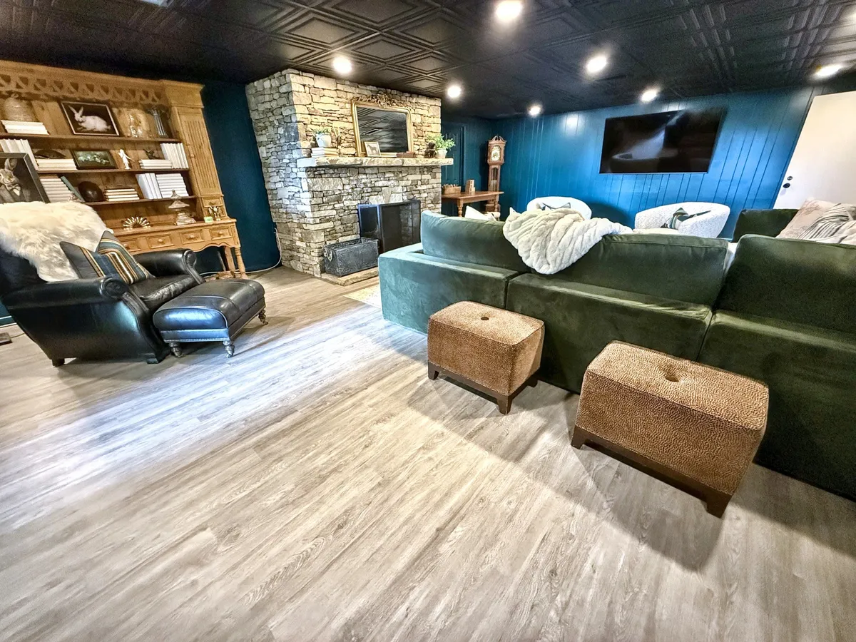 A cozy, well-furnished living area featuring a stone fireplace, a green sectional sofa, a black leather armchair, wooden bookshelves, and a large TV. The space exudes warmth, comfort, and is ideally set up for group movie marathons or game nights.