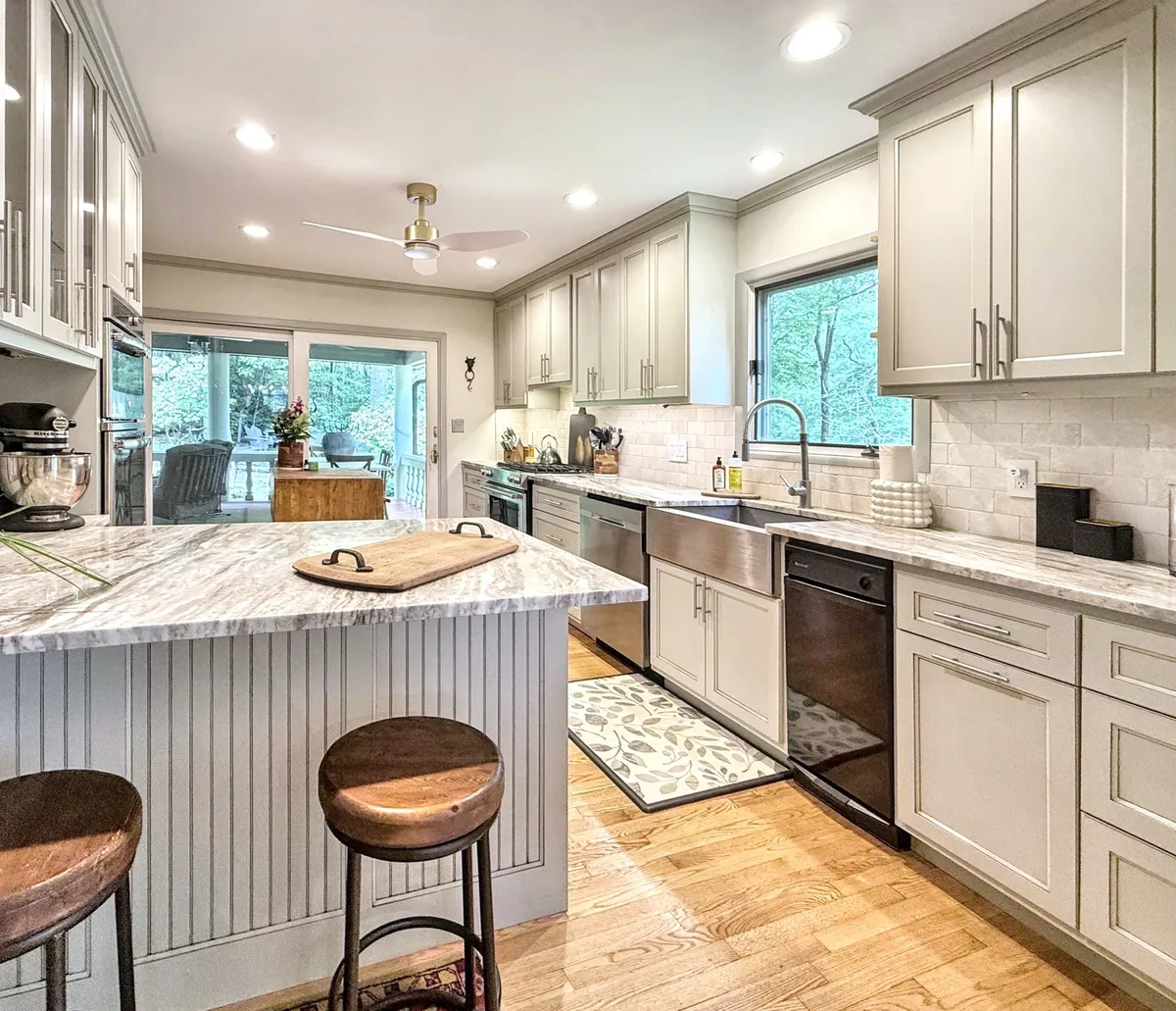 A warmly lit kitchen with a large island countertop, modern appliances, a window offering natural light, wooden floors, and a decorative rug. The inviting space is designed for both functional cooking and friendly gatherings.