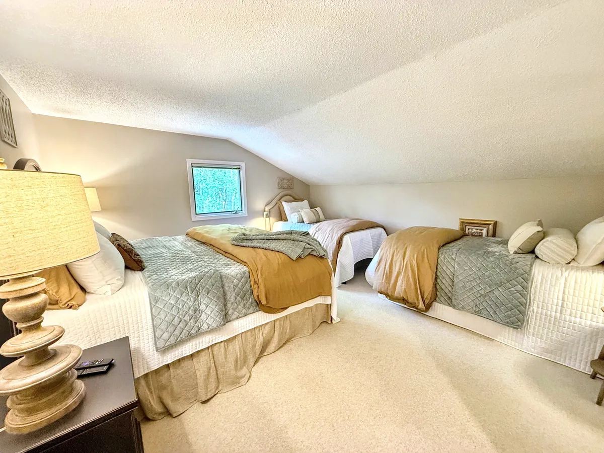 A bright and inviting loft space featuring three beds, a table lamp, and a window that lets in natural light. The arrangement is playful and practical, suited for siblings or cousins to share stories and create memories.
