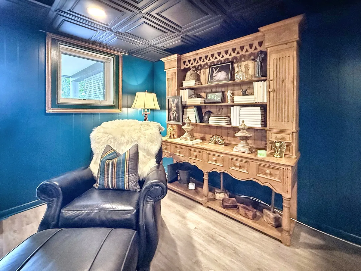 A cozy corner arranged with a black leather chair, a wooden shelf unit, soft lamp lighting, and framed artwork provides an intimate space for reading or reflection. The mood is comfortable and quiet, ideal for personal retreats.
