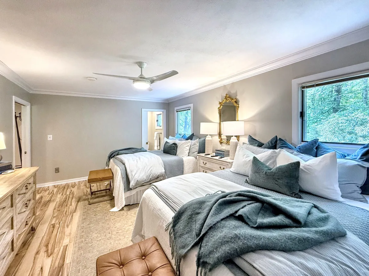 A spacious bedroom with two beds adorned with decorative pillows, a stylish dressing table with lamps, large windows that frame views of greenery, and a decorative mirror. The space feels comfortable, airy, and ideal for sharing.