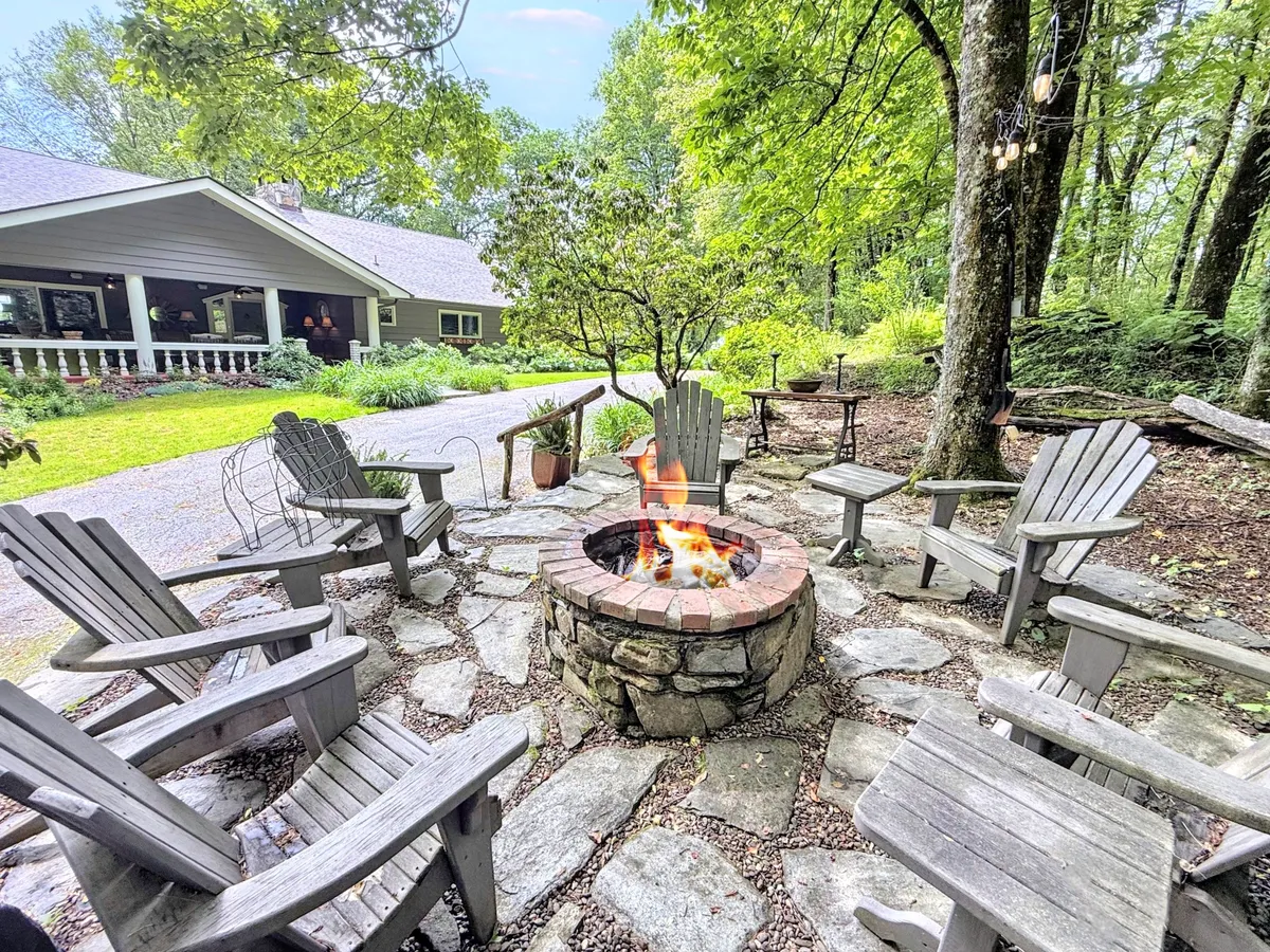 An atmospheric outdoor fire pit scene showing Adirondack chairs arranged in a circle, surrounded by trees, with the house visible in the background. The space is designed for social gatherings, storytelling, and stargazing under the night sky.