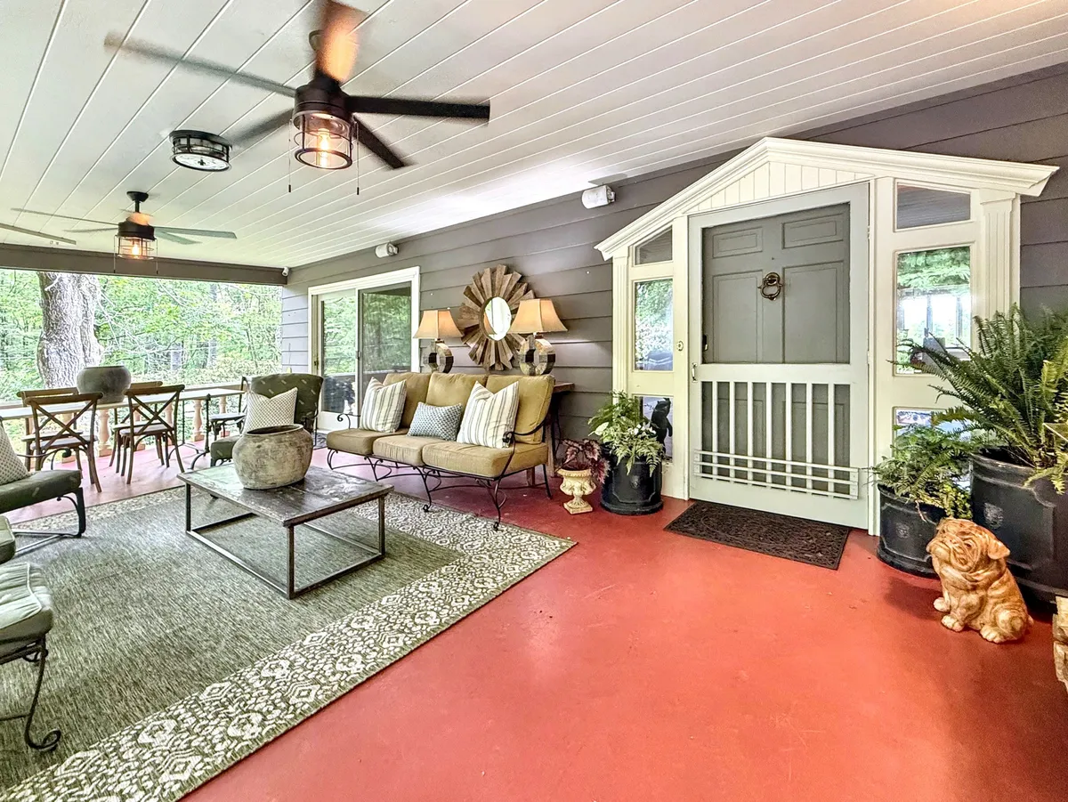 A sunroom or covered deck area furnished with a comfortable sofa, decorative plants, a wooden dining table, and a round mirror. The space is filled with natural light and offers views outside, making it ideal for morning coffee, casual gatherings, or quiet moments of reflection.