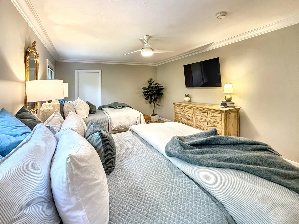 A tranquil bedroom featuring a king-sized bed with layered bedding, a decorative mirror above a dresser, a television mounted on the wall, a nightstand with a lamp, and a vibrant indoor plant. The natural light and soft color palette create a cozy and restful environment.
